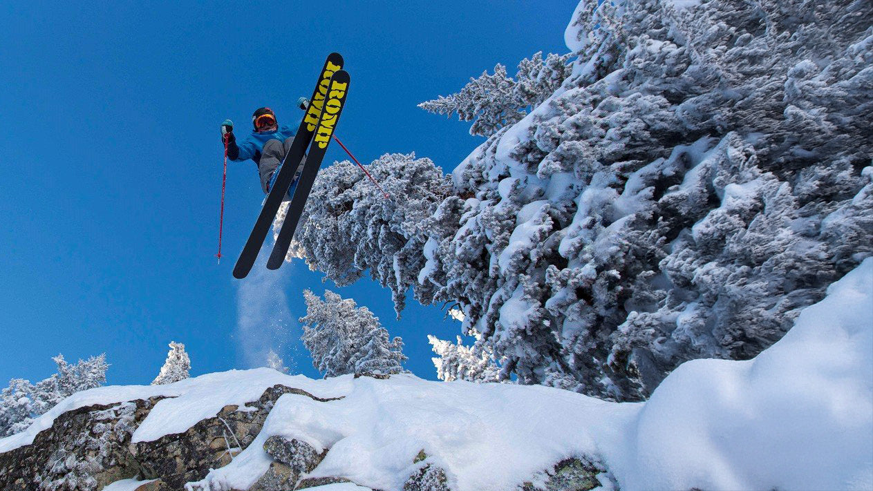 Skiier jumping over the camera with blue sky and pine trees